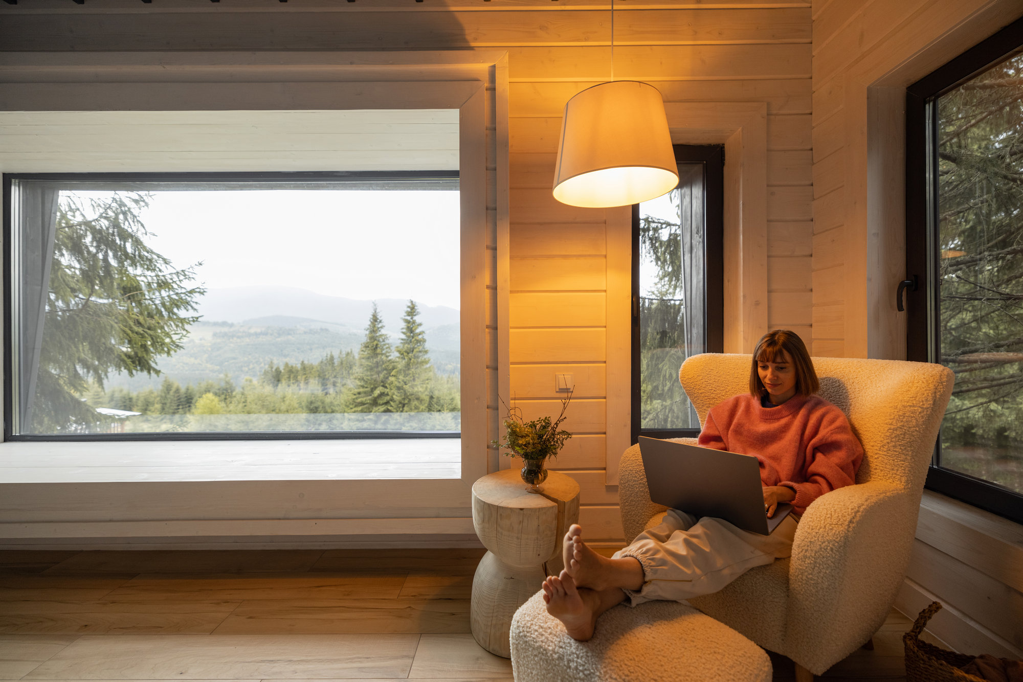 Woman works on laptop while sitting on comfotable sofa in house in mountains. Remote work from cozy place and escaping to nature concept